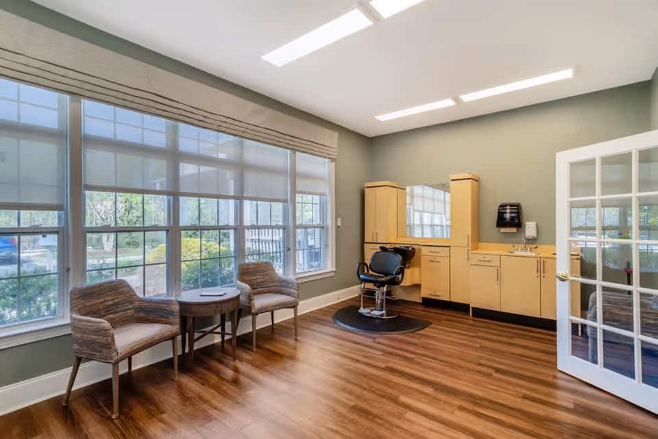 A bright room with large windows covered by roller shades, two upholstered chairs with a small round wooden table between them, and a salon-style chair in front of a wall with light wood cabinetry, a mirror, a sink, and a soap dispenser. The floor is wood and the walls are painted green. A glass-paneled door is open on the right side.