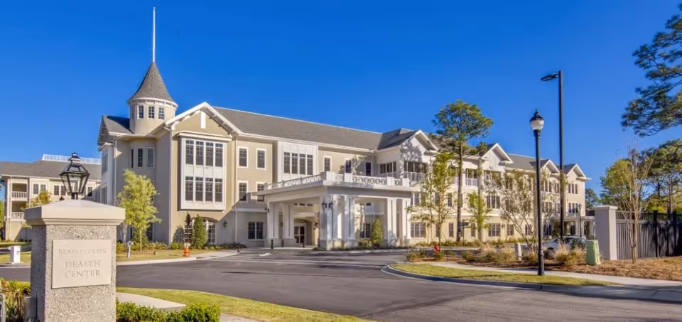 Exterior view of Bradley Creek Health Center at Carolina Bay at Autumn Hall, a large multi-story building with beige siding, white trim, and a covered entrance. The building is surrounded by landscaped greenery and trees under a clear blue sky.