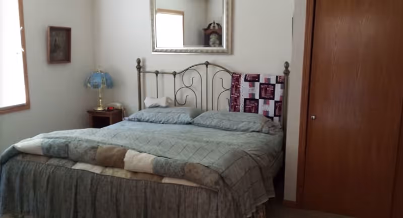 A bedroom with a metal framed bed covered with a green quilt and pillows. There is a wooden nightstand with a blue lamp and a small clock next to the bed. A large mirror hangs on the wall above the bed, and a wooden closet door is visible on the right side of the room.