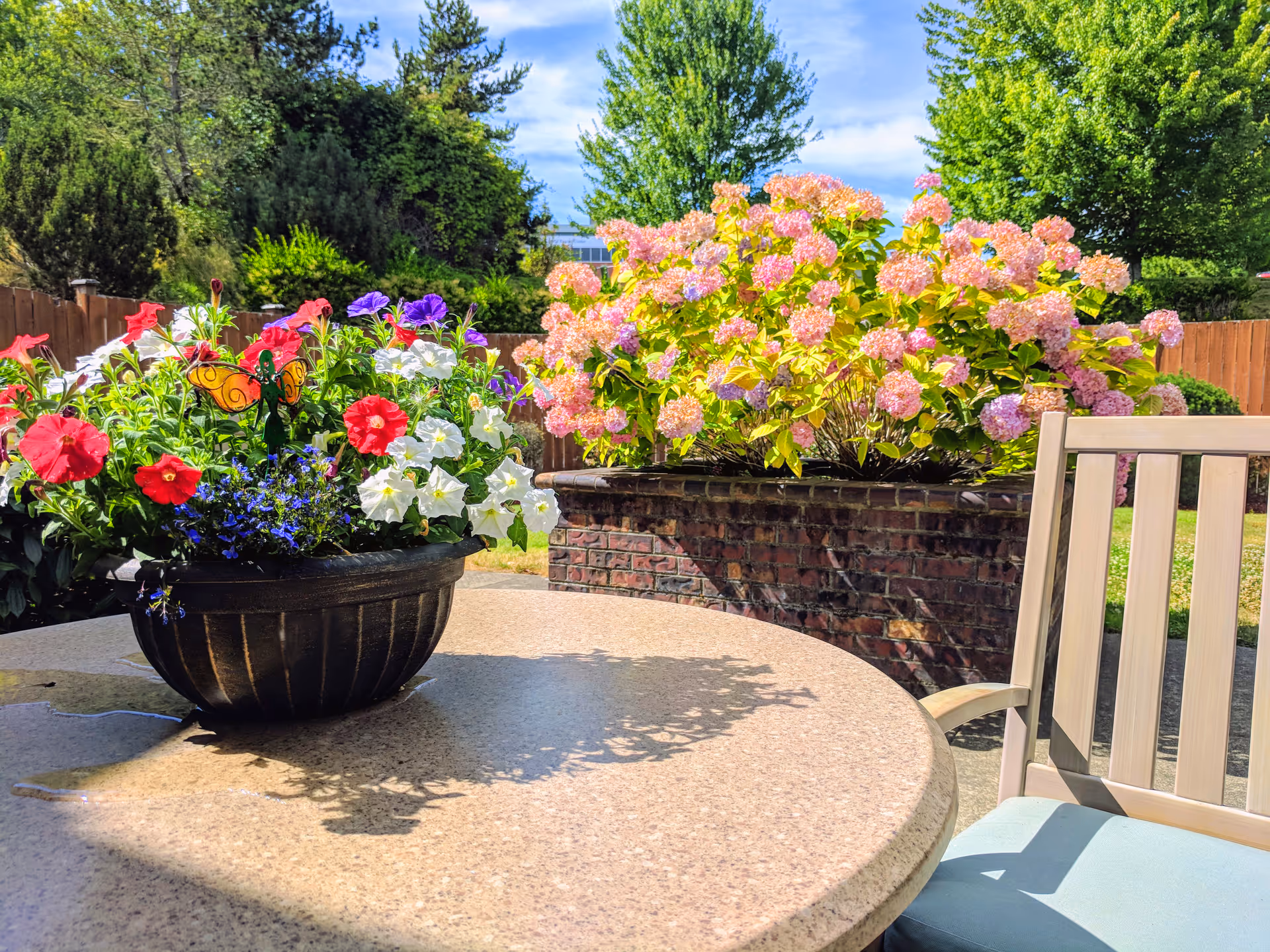 Outdoor patio area with a round stone table featuring a black planter filled with colorful flowers including red, white, purple, and blue blooms. A decorative butterfly is placed among the flowers. In the background, there is a brick planter with pink hydrangea flowers and green trees under a blue sky with some clouds. A beige chair with a light blue cushion is partially visible on the right side of the table.