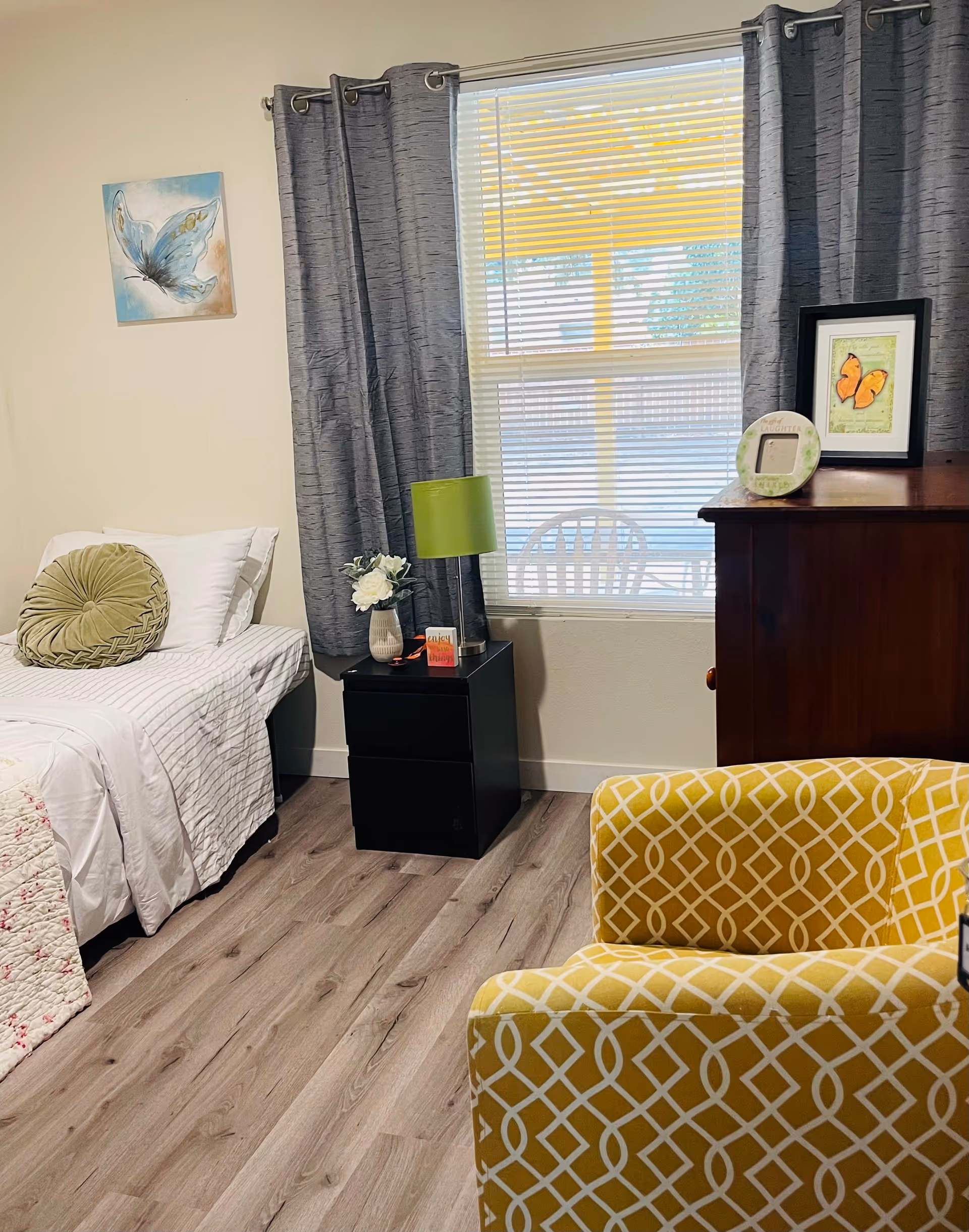 A cozy bedroom with a single bed featuring white and striped bedding and a round green pillow. Next to the bed is a black nightstand with a green lamp, a small vase with white flowers, and a decorative item. A window with gray curtains and blinds lets in natural light. A wooden dresser with framed butterfly artwork and a small round frame sits beside the window. In the foreground, there is a yellow armchair with a white geometric pattern. The room has light wood flooring and a butterfly painting on the wall.