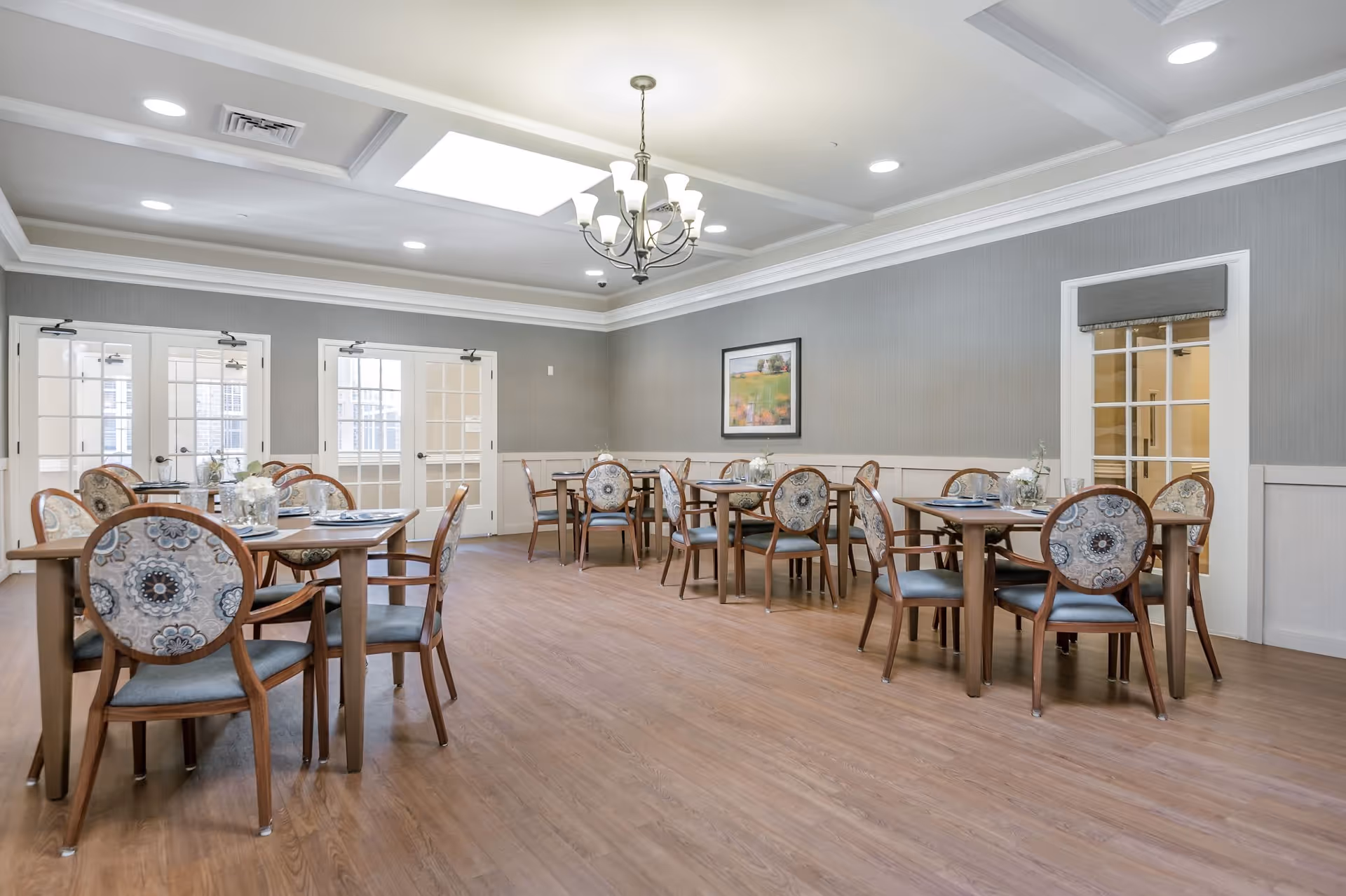 A bright and spacious dining room with several wooden tables and chairs arranged neatly. The chairs have patterned upholstery and the room features a chandelier, recessed lighting, and a skylight. The walls are painted gray with white trim, and there are multiple glass-paneled doors and a framed picture on the wall.