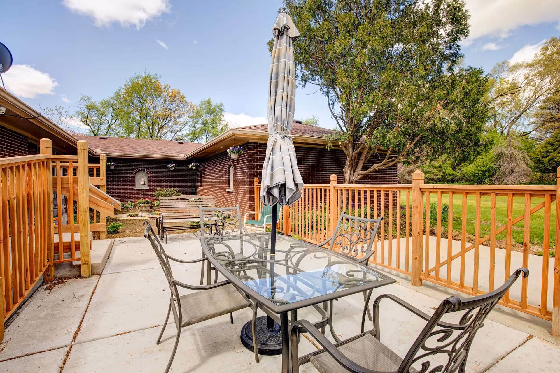 Outdoor patio area with a glass-top table and four metal chairs with decorative backs. A closed striped umbrella stands in the center of the table. The patio is surrounded by a wooden fence and has a concrete floor. In the background, there is a brick building with hanging flower pots and a wooden bench. Trees and green grass are visible beyond the patio.