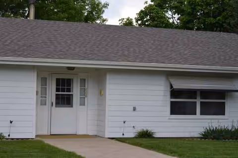 White single-story facility exterior with a front entrance, window, lawn, and sidewalk.