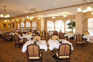 A spacious dining room with multiple tables covered in white tablecloths, each set with plates, glasses, and napkins. The room features large windows with beige curtains, patterned carpet, and several ceiling light fixtures providing warm lighting. Chairs with striped upholstery surround the tables, and a potted plant is visible near the windows.