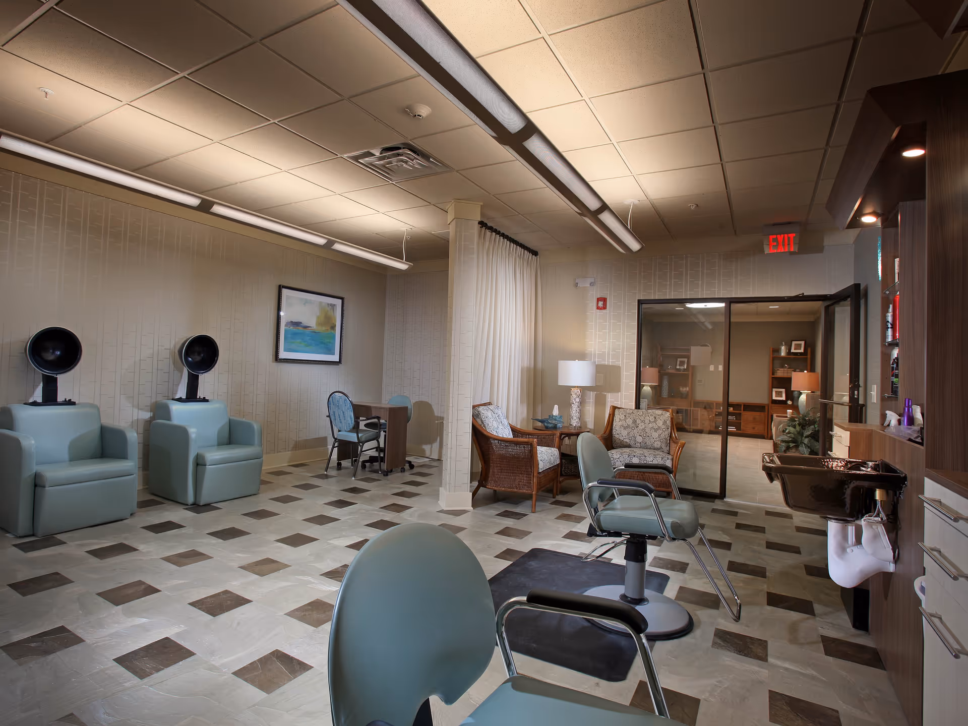 Interior of a senior living facility salon area with two hair drying chairs on the left, a manicure table with two chairs in the back, and two salon chairs in the foreground near a sink. There are also two wicker armchairs with cushions and a side table with a lamp near a glass door leading to another room.