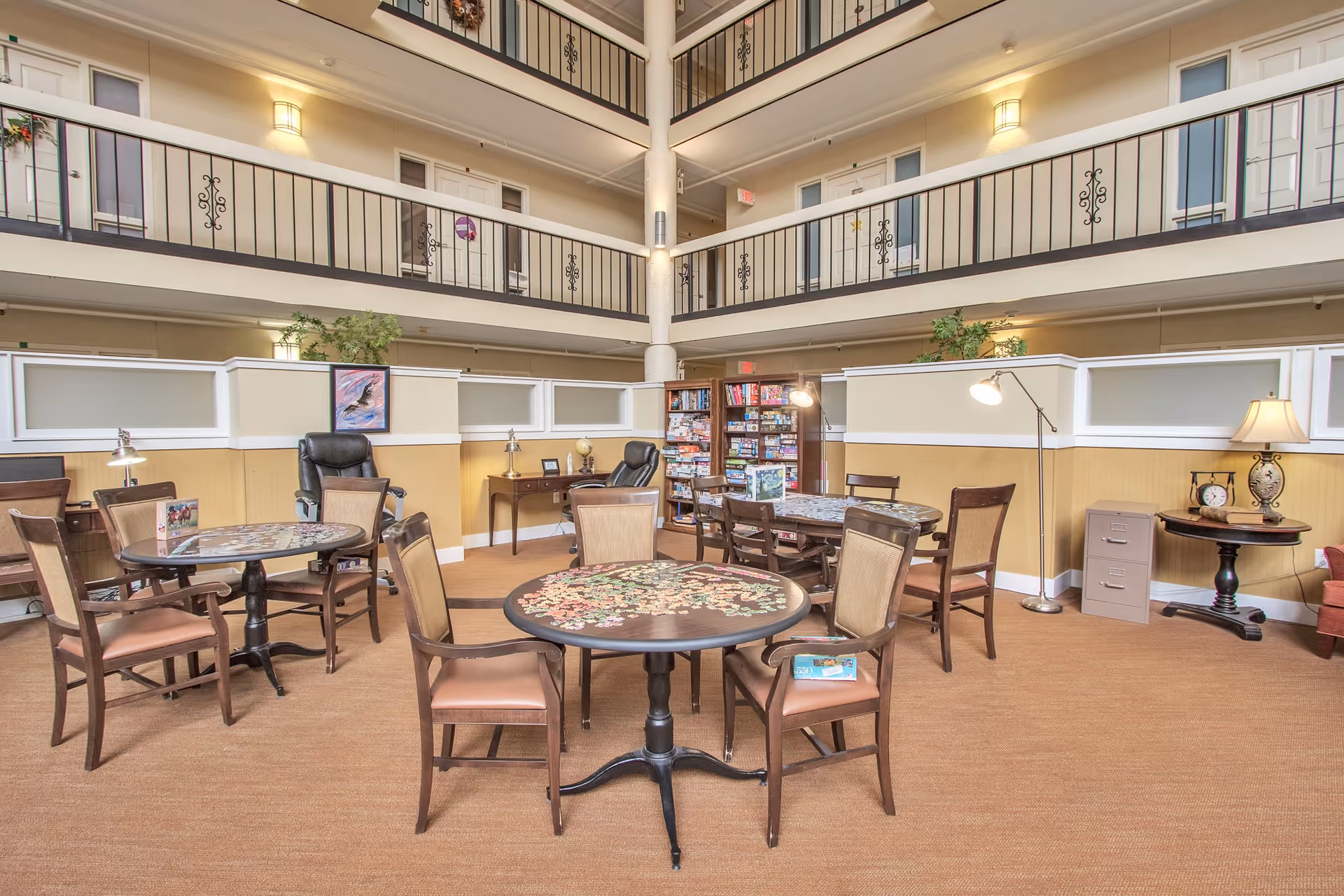 A spacious common area in a senior living facility with multiple round tables and chairs arranged for socializing or activities. The room has a high ceiling with two upper floors visible, each with railings and doors to rooms. There are lamps, a bookshelf filled with board games, and decorative plants, creating a welcoming atmosphere.