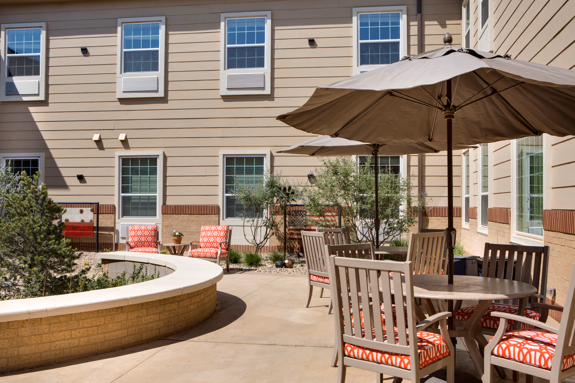 Outdoor courtyard area at The Legacy at South Plains featuring beige building walls with multiple windows, a curved brick planter with greenery, patio tables with umbrellas, and chairs with orange patterned cushions.
