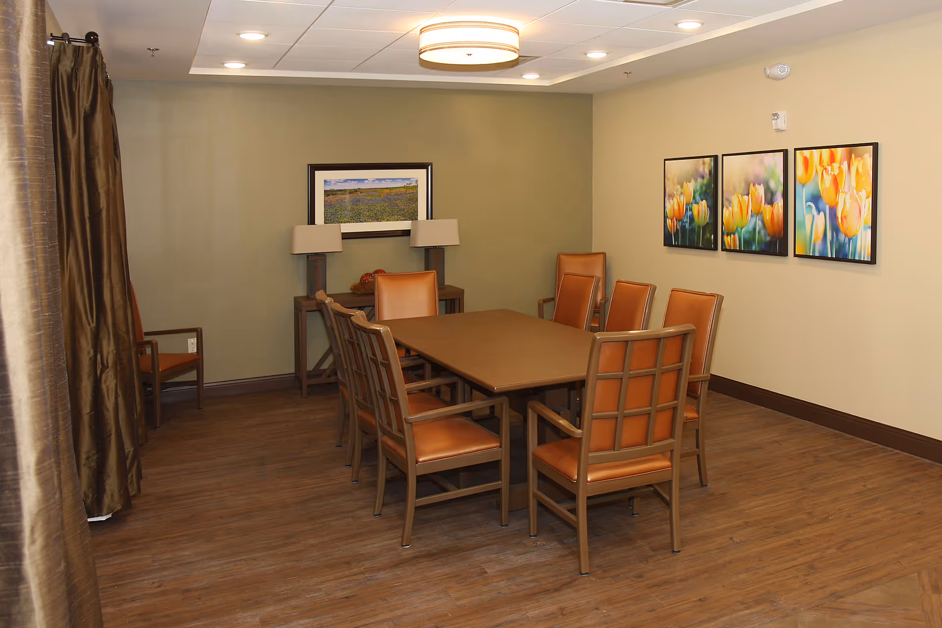 A dining room with a rectangular wooden table surrounded by eight brown cushioned chairs. The room has wooden flooring, beige walls, and three framed pictures of yellow and orange tulips on one wall. There is a small console table with two lamps and a framed landscape photo on the far wall. Brown curtains are partially visible on the left side.