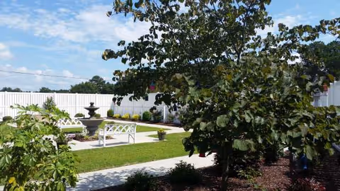 Outdoor courtyard with a fountain, benches, and leafy trees beside a white fence.