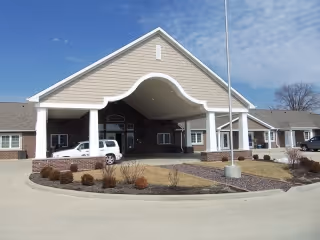 Front exterior view of a single-story senior living facility building with a covered entrance, a white vehicle parked under the entrance canopy, landscaped bushes, and a flagpole in front.