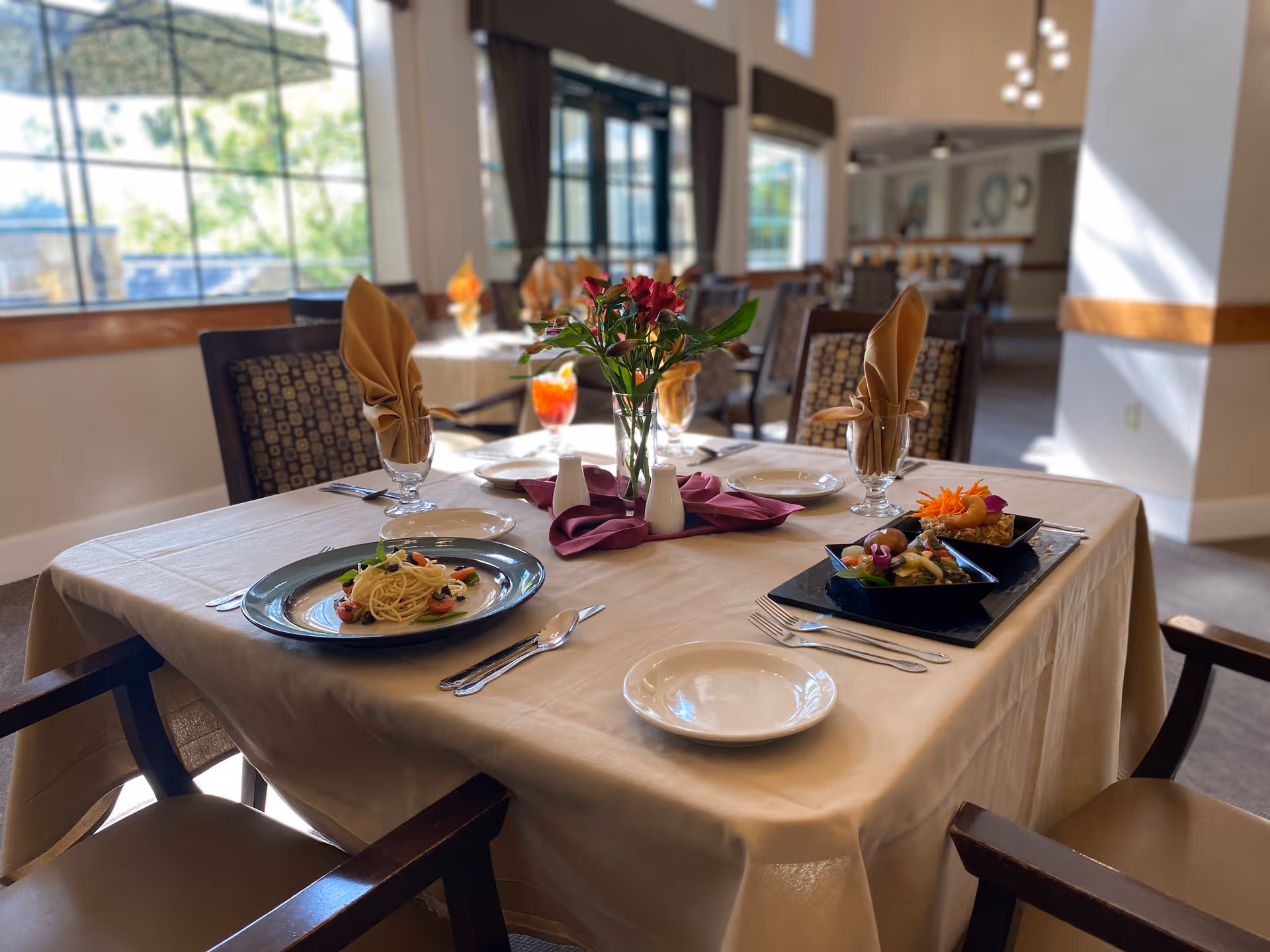 A dining table set for four in a bright dining room with large windows. The table is covered with a beige tablecloth and has plates of food, glasses with folded napkins, silverware, and a centerpiece with flowers. The room has multiple tables and chairs arranged for dining.
