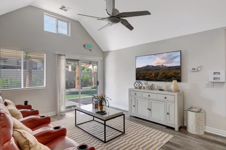 A bright living room with vaulted ceilings featuring three red leather recliners with beige pillows, a rectangular coffee table with a plant and remote controls, a white sideboard with decorative items, and a large flat-screen TV mounted on the wall displaying a scenic landscape. There are windows with blinds and a sliding glass door leading to an outdoor area with greenery.