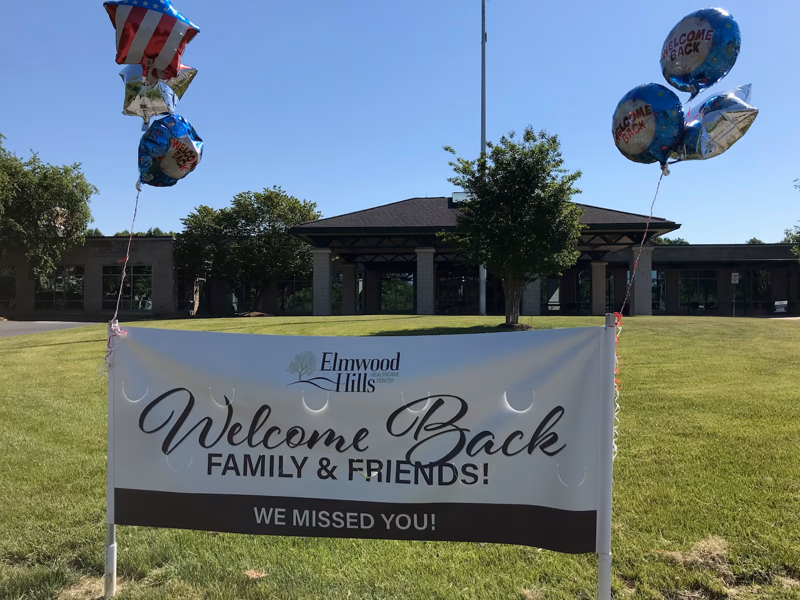 Outdoor view of Elmwood Hills Healthcare Center with a large white banner in the foreground that reads 'Welcome Back Family & Friends! We Missed You!' The banner is decorated with balloons, some with American flag designs and others with 'Welcome Back' text. The building is surrounded by green grass and trees under a clear blue sky.