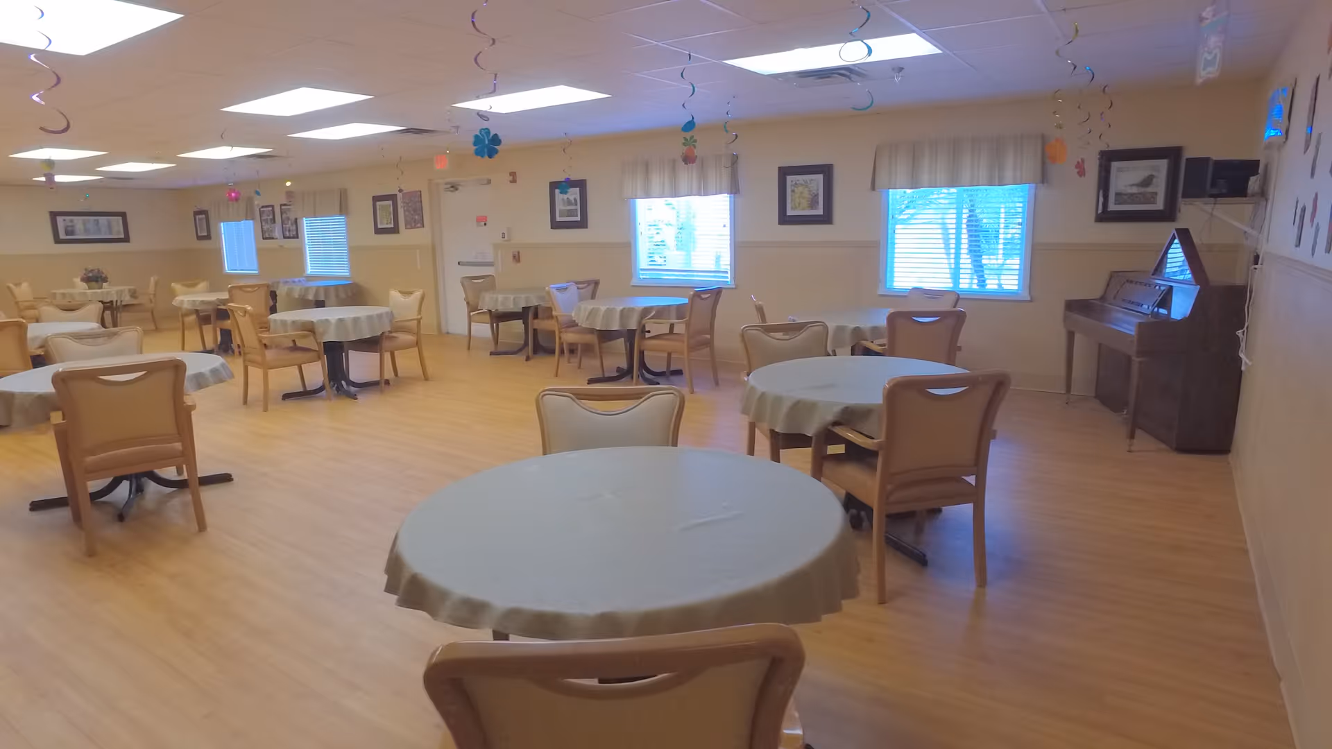 A spacious dining room with multiple round tables covered with light-colored tablecloths, each surrounded by beige chairs. The room has wooden flooring, several windows with blinds and valances, framed pictures on the walls, and a piano in the corner. Colorful hanging decorations are suspended from the ceiling.