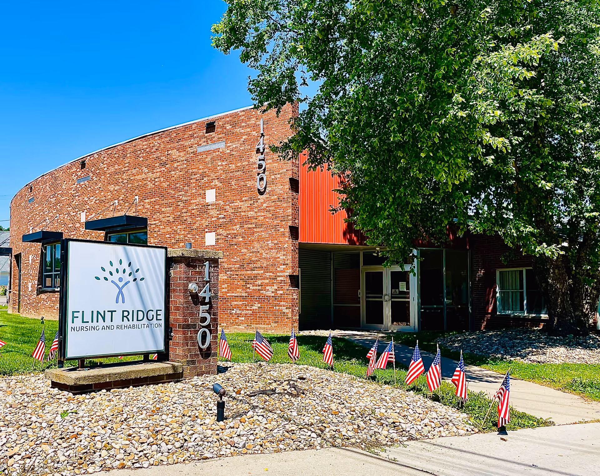 Exterior view of Flint Ridge Nursing and Rehabilitation building with a brick facade, a large sign displaying the facility's name and logo, a row of small American flags planted in the ground, a tree providing shade, and a clear blue sky.