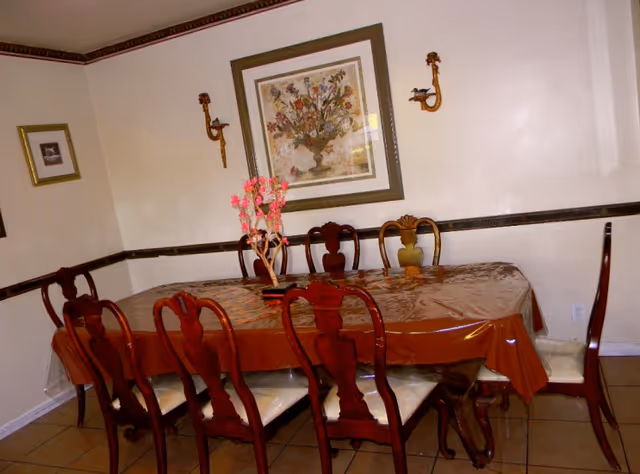 Dining room with a long wooden table covered by a plastic tablecloth, eight matching chairs, a floral centerpiece, and framed artwork on the wall.