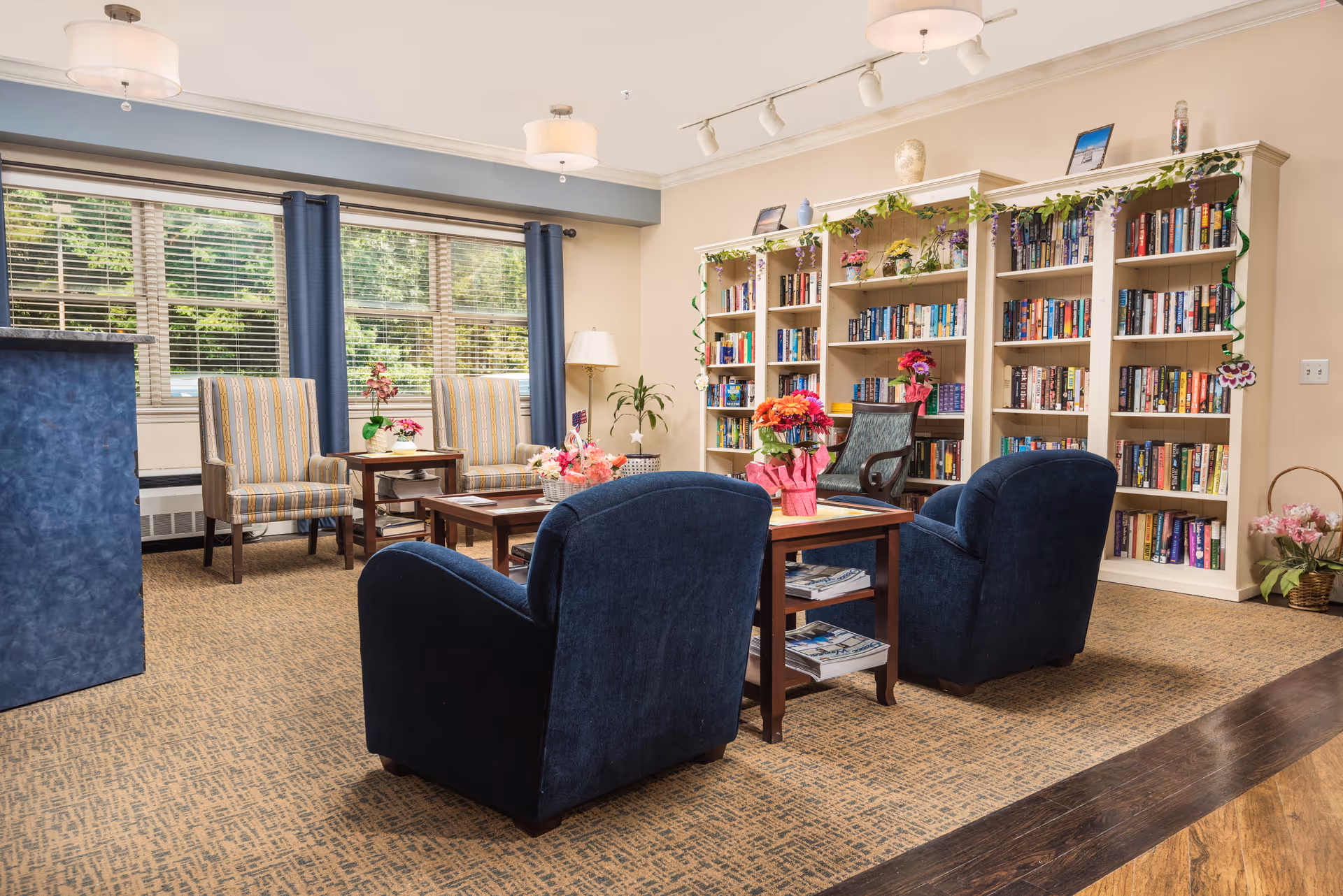 A cozy reading and sitting area in a senior living facility with two blue armchairs, two striped armchairs, a wooden coffee table with flowers, and a large bookshelf filled with books decorated with green garlands and flowers. Large windows with blue curtains allow natural light to fill the room.