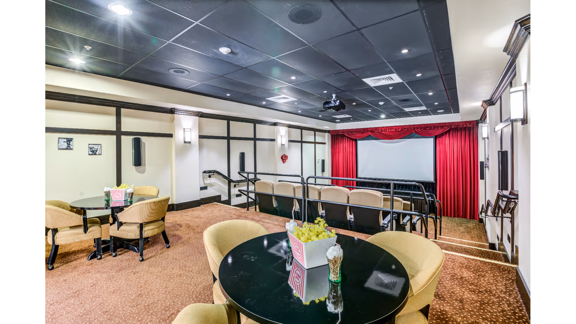 A small theater room with a projector screen framed by red curtains. There are two rows of theater seating with beige chairs and black railings. In the foreground, there are round tables with beige upholstered chairs, one table has a popcorn container and a decorative centerpiece. The walls are light-colored with dark wood trim and sconces, and the ceiling is black with recessed lighting.