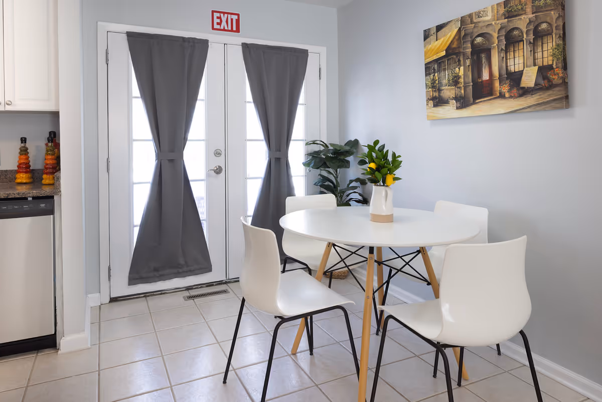 Bright dining area with a round white table and four chairs in front of glass French doors covered by gray curtains and an EXIT sign above.
