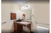 Small accessible bathroom with a vanity and sink under a large mirror, light fixture above, toilet, grab bars, and beige walls.