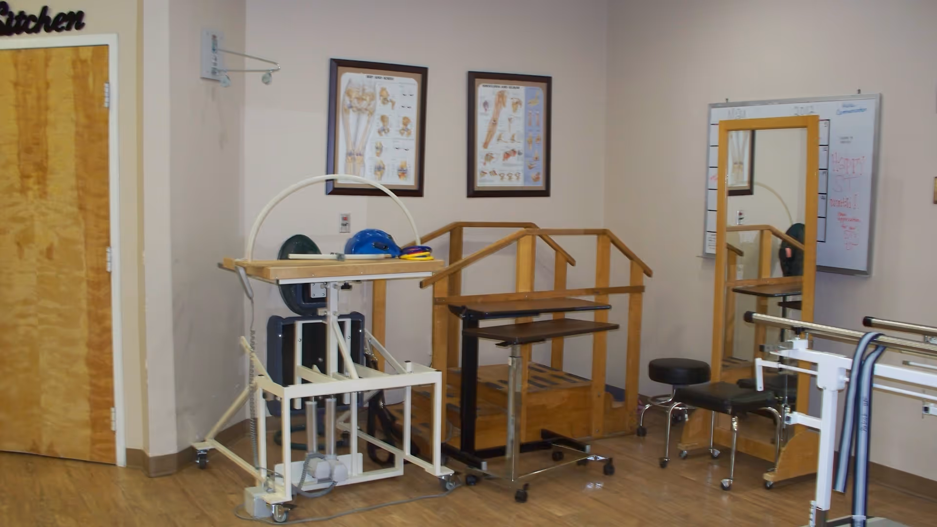 A rehabilitation therapy room with wooden parallel bars, a standing frame device, a rolling table, a mirror, and anatomical charts on the wall. The room has wooden flooring and light-colored walls.
