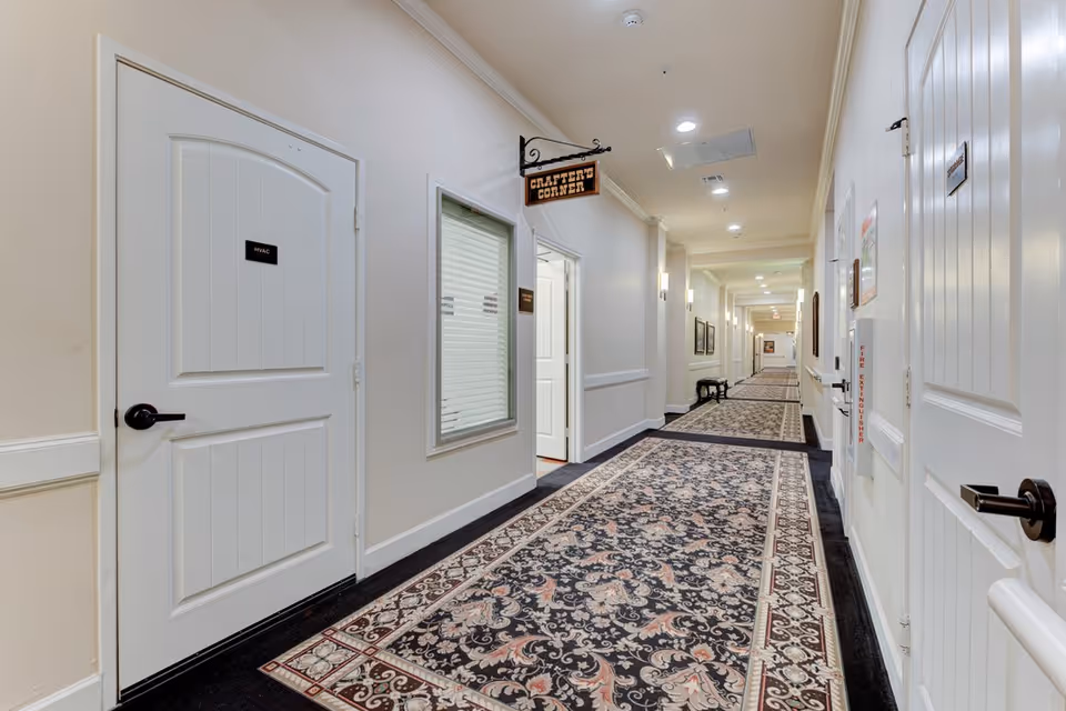A long, well-lit hallway in a senior living facility with patterned carpet, cream-colored walls, and multiple white doors. One door is labeled HVAC, and a sign hanging from the ceiling reads 'Crafters Corner.' There are framed pictures on the walls and a bench along the hallway.