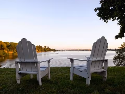 Two white Adirondack chairs on a grassy area facing a calm body of water with trees on the far shore and a clear sky at sunset or sunrise.