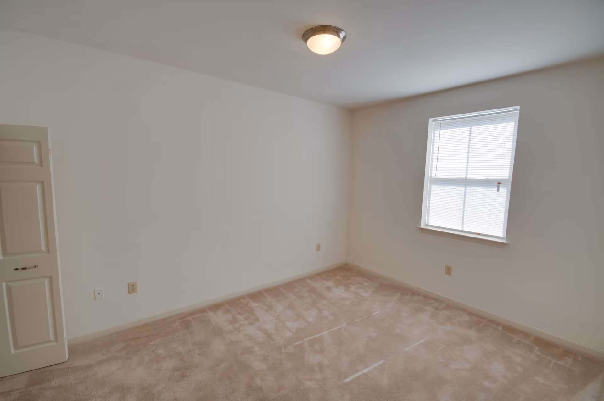 Empty room with beige carpet, white walls, a window with blinds, a ceiling light fixture, and a partially open door.
