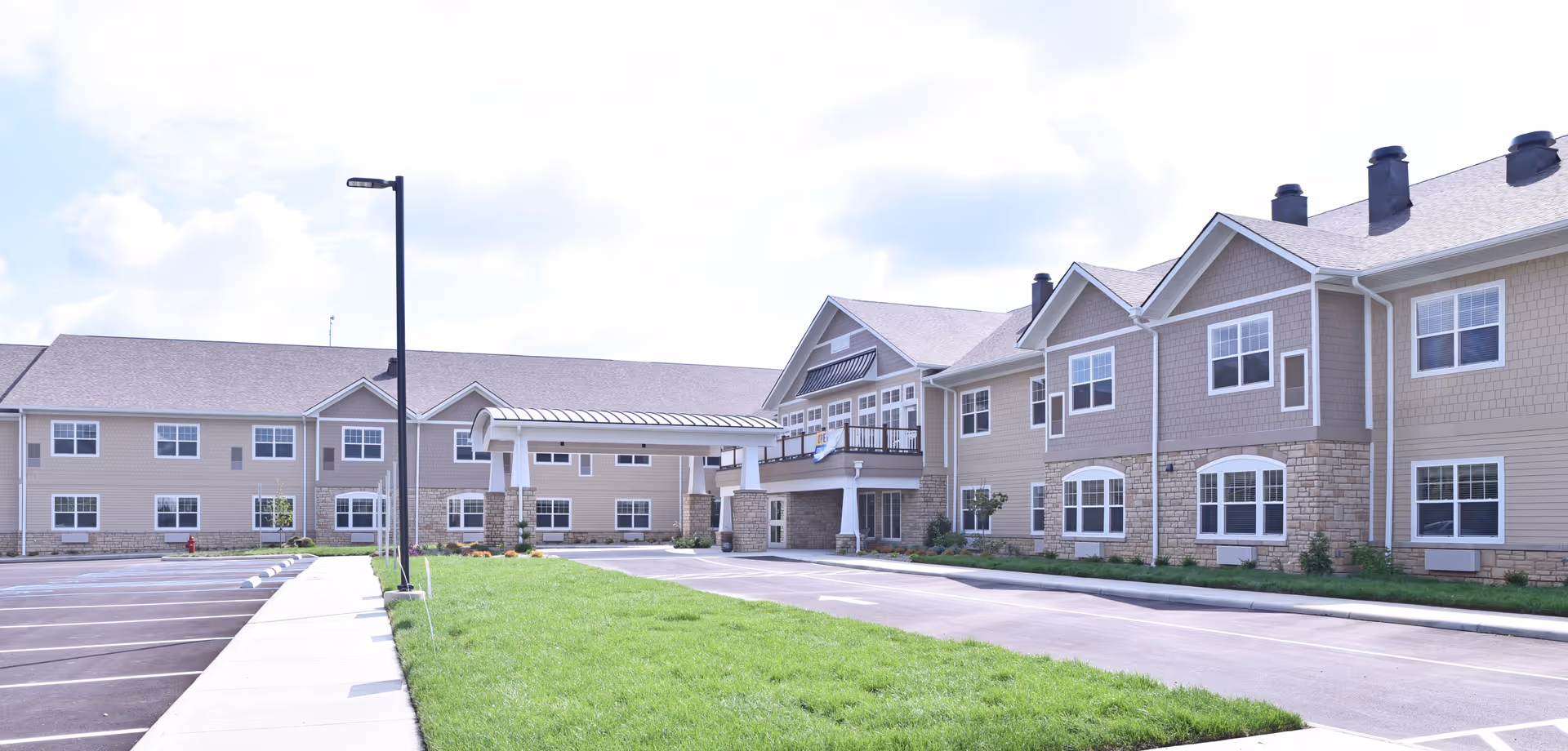 Exterior front view of a two-story senior living facility with a covered porte-cochère, parking lot, and landscaped lawn.
