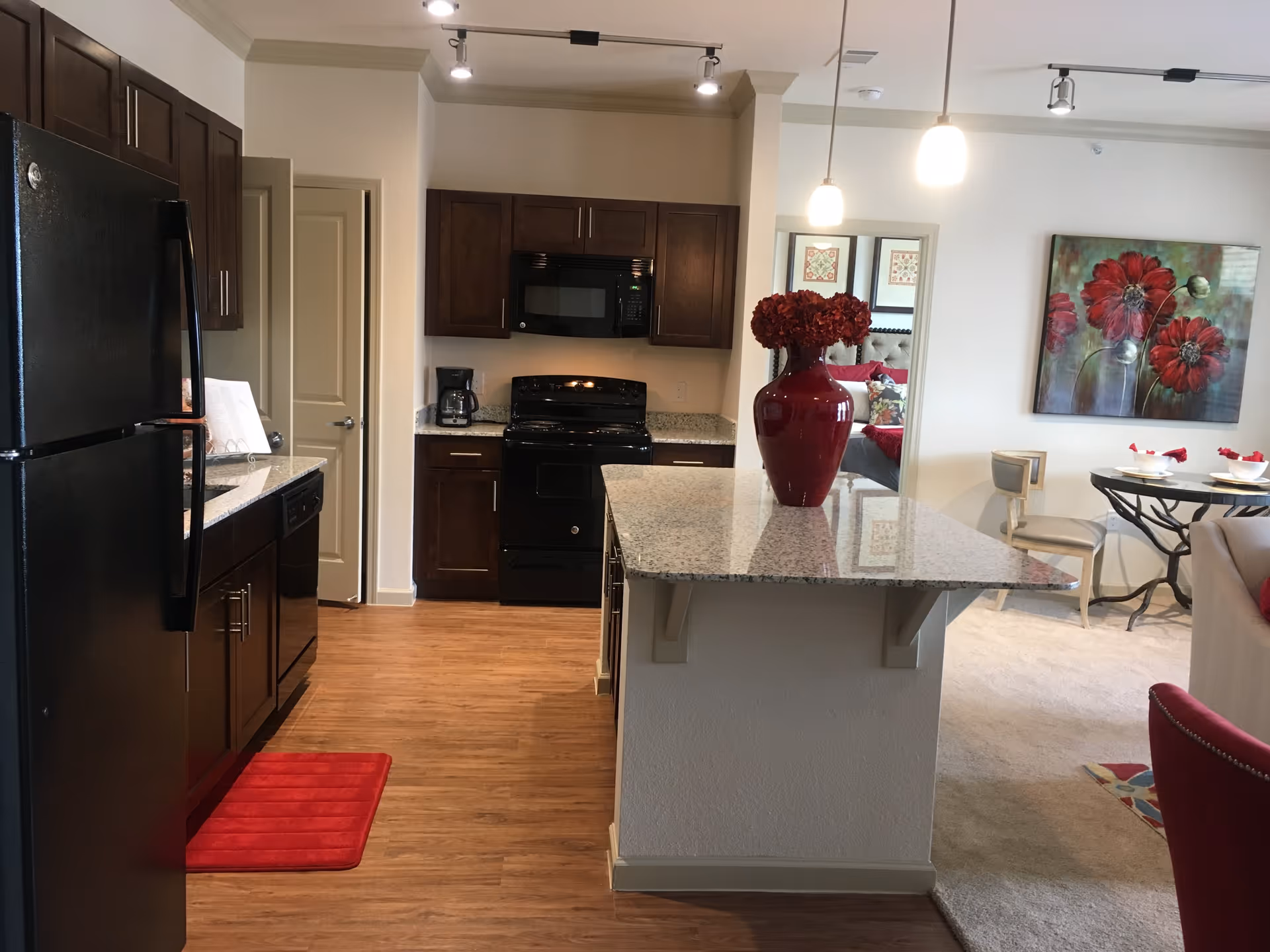 Modern kitchen with dark wood cabinets, black appliances including refrigerator, stove, microwave, and dishwasher. Granite countertop island with a red vase holding red flowers. Adjacent dining area with a round table, chairs, and a large floral painting on the wall. Hardwood floor in the kitchen area and carpet in the dining and living space.