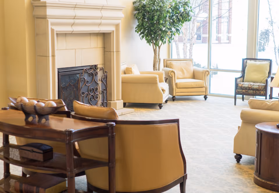 A bright and cozy living room area with beige armchairs and a sofa arranged around a decorative fireplace. There is a wooden table with a bowl of decorative balls and a small box. Large windows let in natural light and show a snowy outdoor scene with trees.