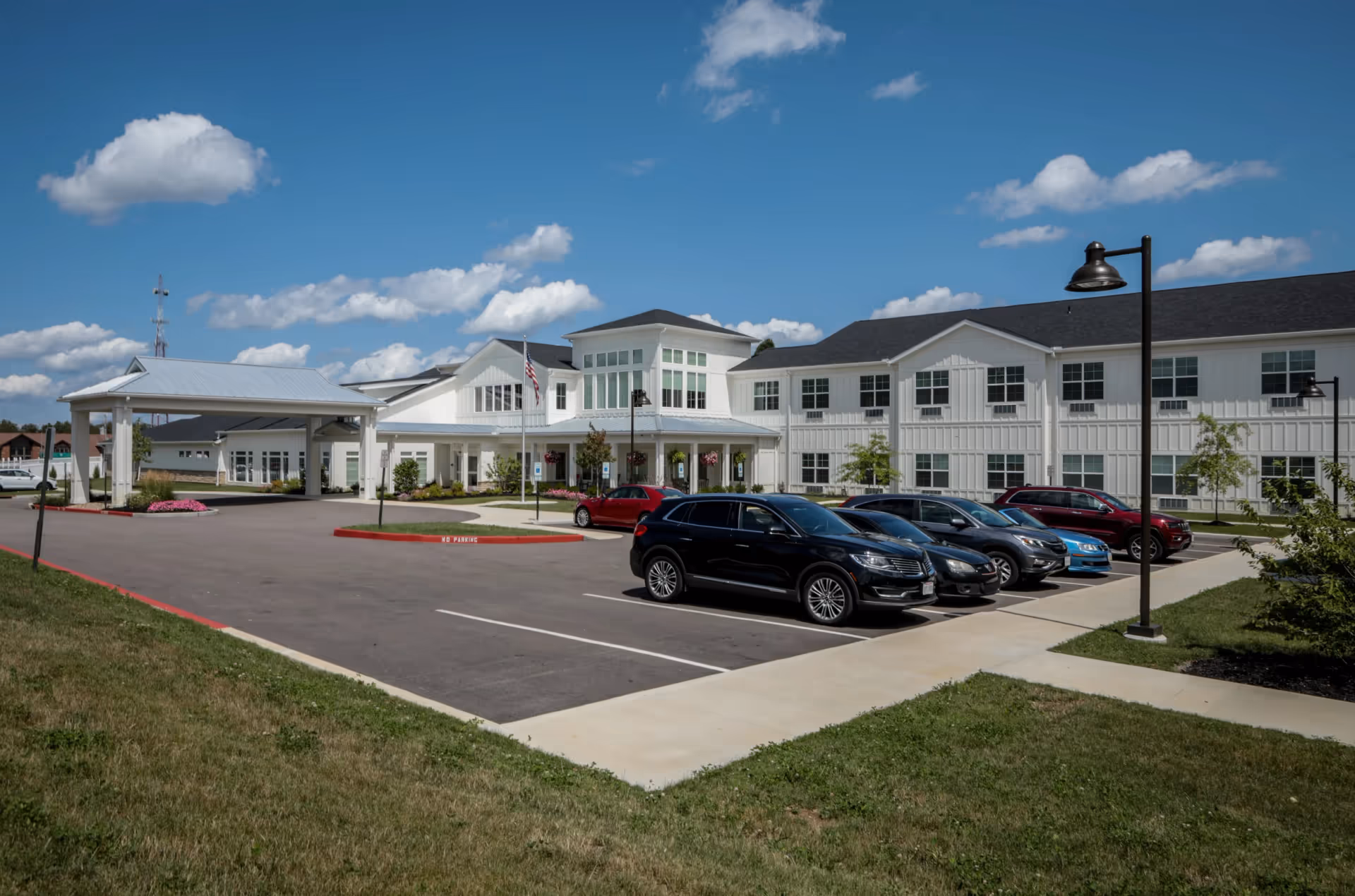 Exterior view of Heritage Crossing senior living facility showing a large white building with many windows, a covered entrance, several parked cars, a flagpole with an American flag, and a clear blue sky with scattered clouds.