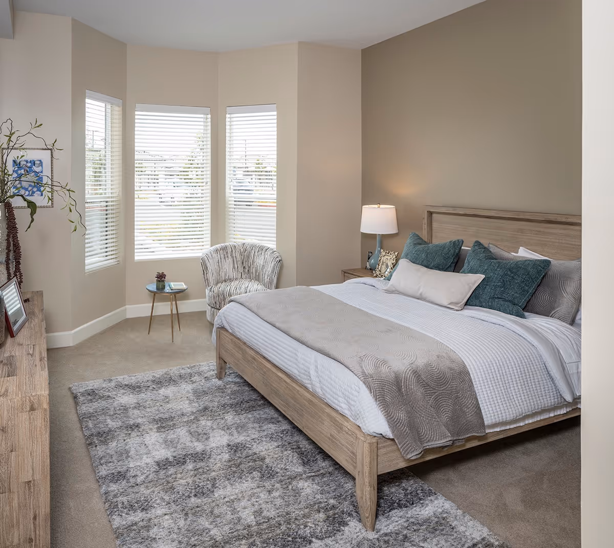 A cozy bedroom with a wooden bed frame, neatly made bed with white and beige bedding, and several green and beige pillows. There is a bedside table with a lamp and picture frames. A patterned armchair and a small round table with a plant and book are placed near three windows with white blinds. A large gray and beige rug covers part of the carpeted floor.