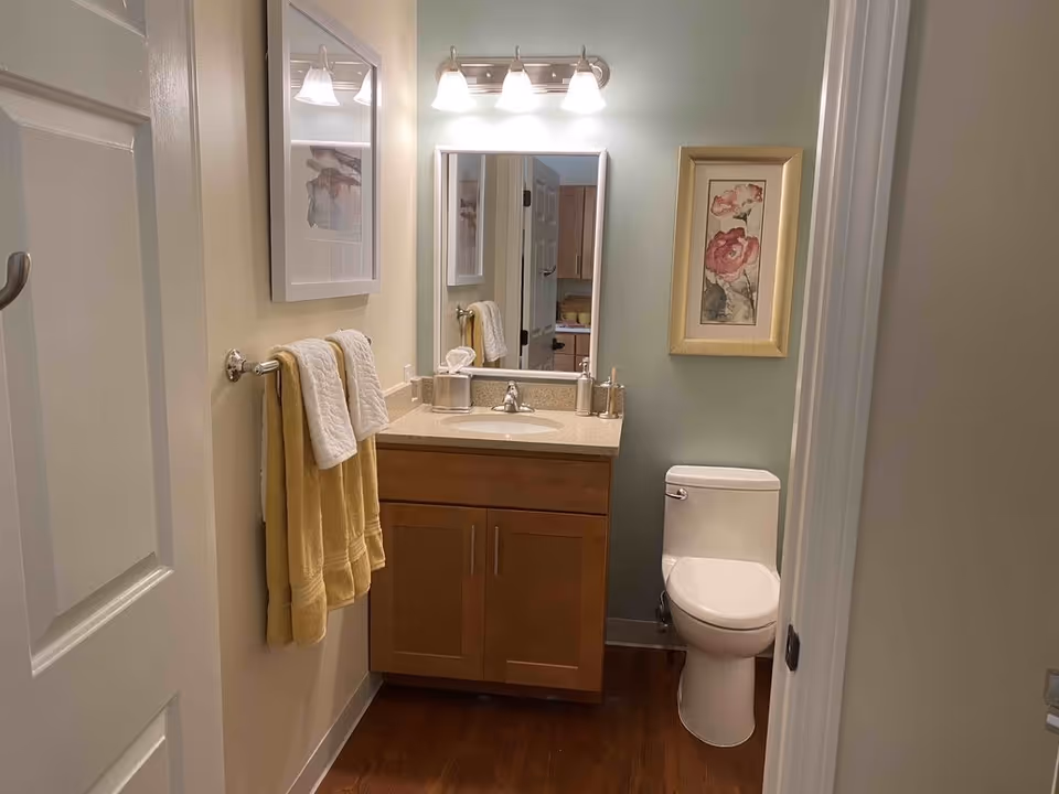 A small bathroom with a wooden vanity cabinet topped with a beige countertop and a sink. Above the sink is a rectangular mirror with three light fixtures. To the right of the vanity is a white toilet. On the left wall, there are two yellow towels hanging on a towel rack beneath a framed picture. Another framed floral artwork hangs on the wall above the toilet. The floor is wooden, and the walls are painted in light colors.
