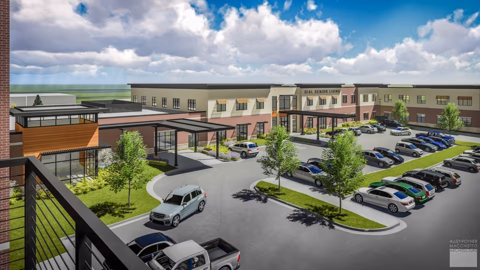 Exterior view of Silvercrest at College View Senior Living facility showing a modern two-story building with multiple windows, a covered entrance, landscaped greenery, and a parking lot with several cars parked. The sky is partly cloudy with blue patches.