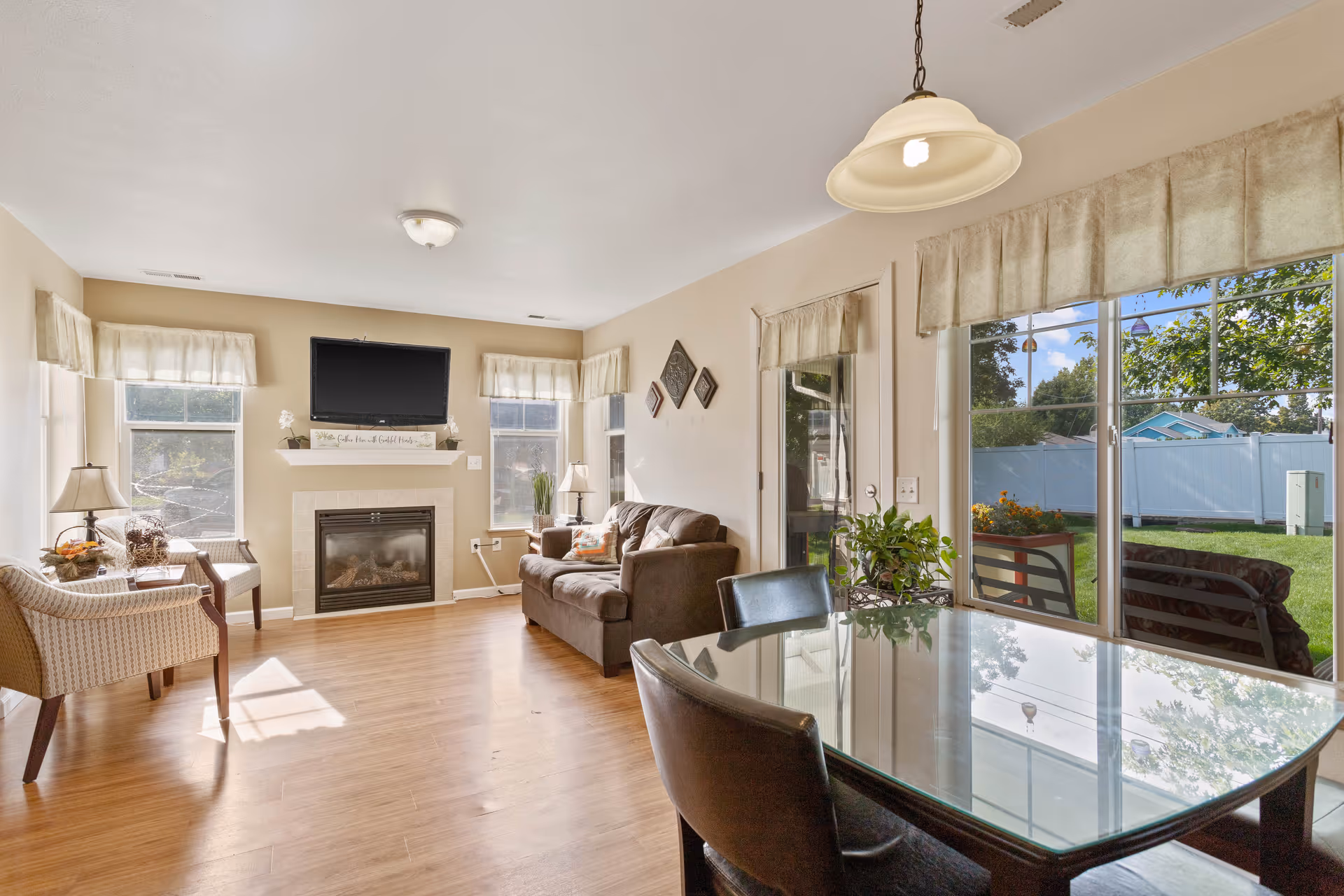 A bright living room and dining area with wooden flooring. The living room features a brown sofa, two beige armchairs, a fireplace with a mounted flat-screen TV above it, and windows with light curtains allowing natural light to enter. The dining area has a glass-top table with four black chairs and a hanging light fixture above it. Large sliding glass doors provide a view of a green outdoor space with a white fence and trees.