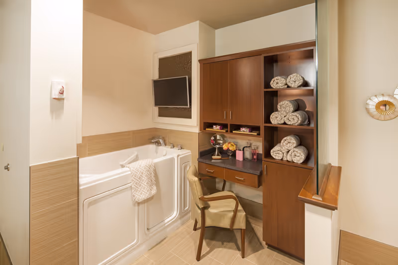 A bathroom featuring a walk-in bathtub with a towel hanging on the side, a mounted flat-screen TV above the tub, and a wooden vanity with a chair. The vanity has rolled towels on open shelves, a mirror, and various toiletries on the countertop. The walls and floor have beige tiles, and there is a decorative wall clock on the right.