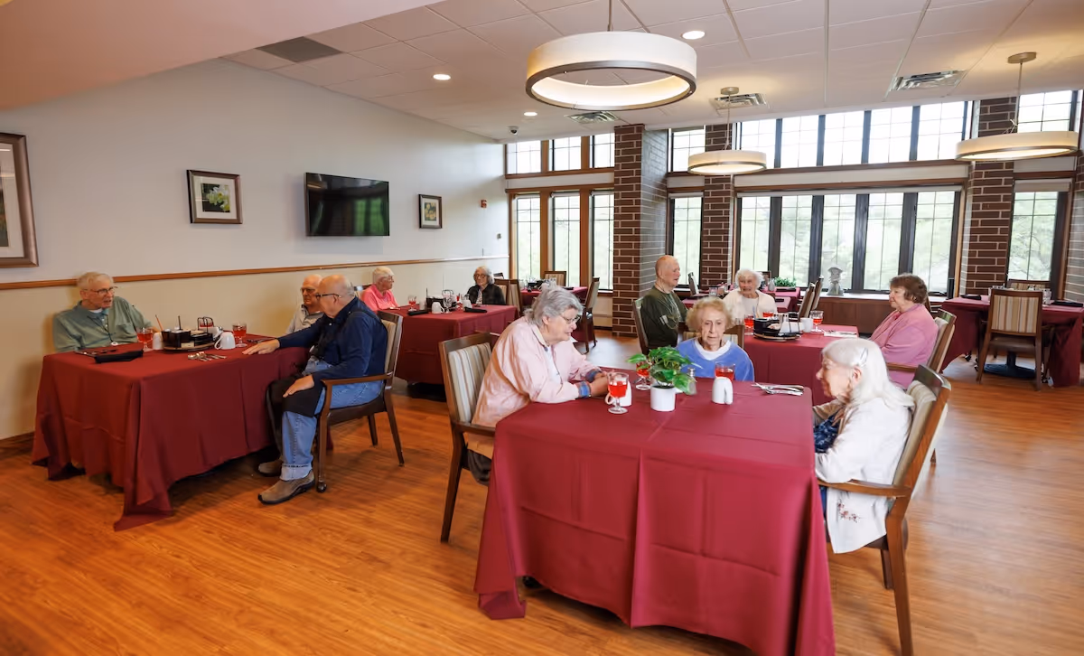 A dining room in a senior living facility with several elderly residents seated at tables covered with red tablecloths. The room has large windows letting in natural light, wooden floors, and modern circular ceiling lights. Some residents are engaged in conversation while others sit quietly.