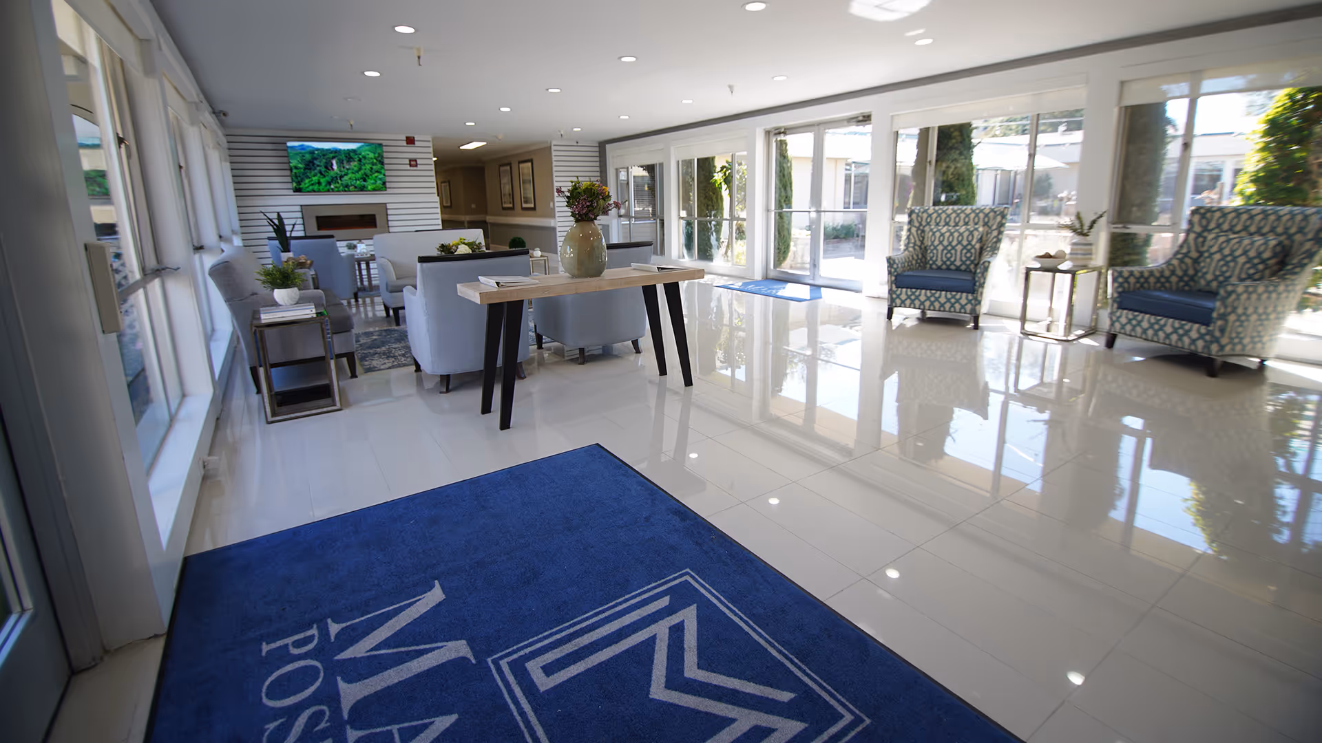 Bright and spacious lobby area of Marin Post Acute featuring a blue entrance mat with the facility's logo, modern seating with armchairs and sofas, a wooden table with a vase of flowers, large windows allowing natural light, and a wall-mounted TV above a fireplace.