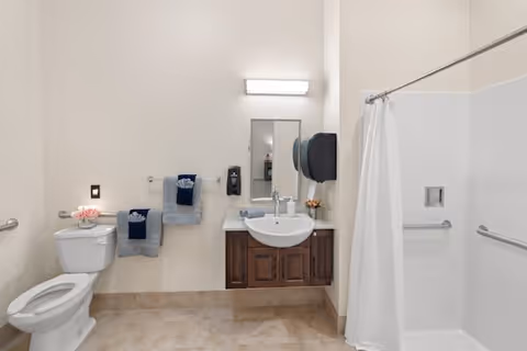 A clean and accessible bathroom featuring a toilet with a flower vase on top, a wall-mounted sink with a mirror and light above it, two towel racks with folded towels, a soap dispenser, a paper towel dispenser, and a shower area with a white curtain and grab bars.