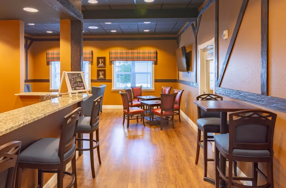 Interior view of a senior living facility dining area with wooden flooring, orange walls, and dark wood trim. The room features a granite countertop bar with high chairs on the left, a small table with four red cushioned chairs in the center near two windows with plaid valances, and a high table with two chairs on the right. A small chalkboard on the bar displays the words 'Royal Spritzer'. A flat-screen TV is mounted on the right wall.