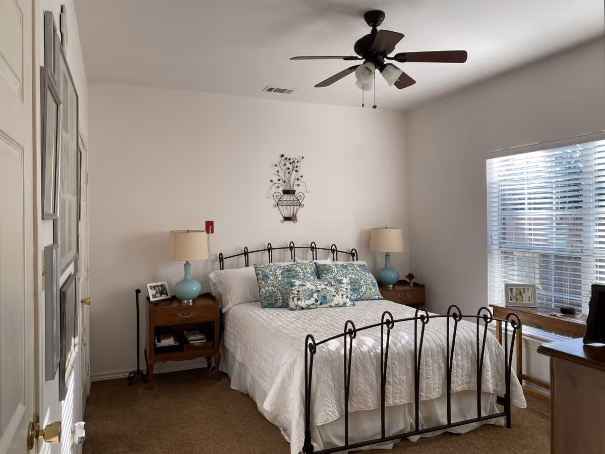 Sunlit bedroom with a black metal bed frame, white quilt and patterned pillows, bedside tables with lamps, a ceiling fan, and a window with blinds.