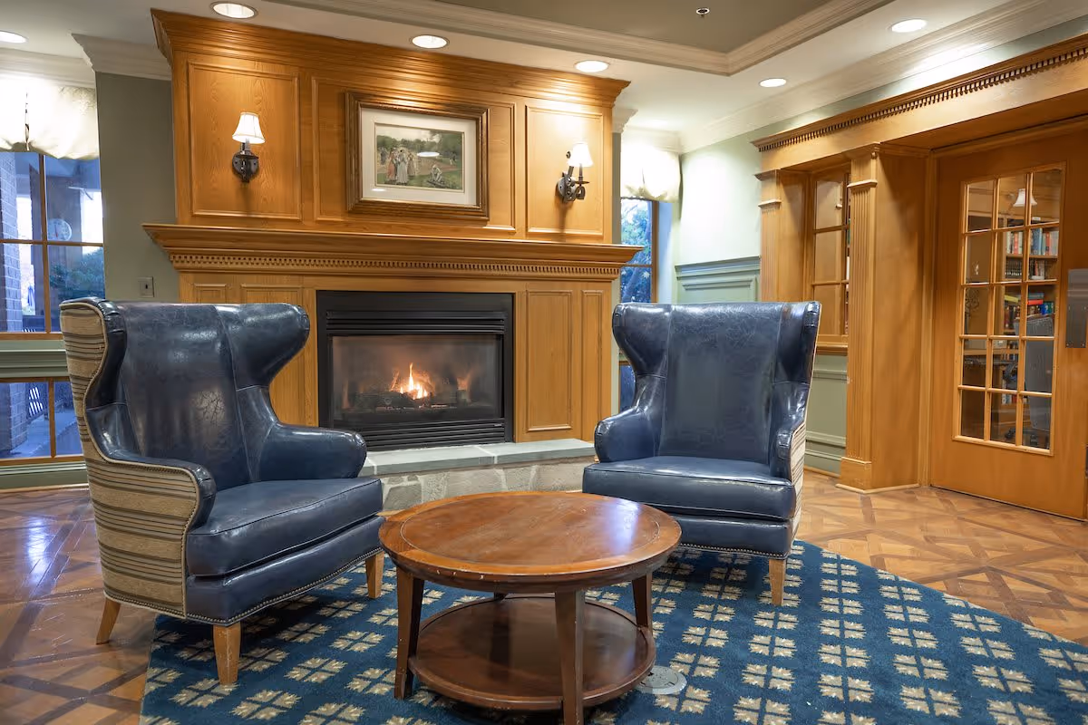 A cozy sitting area with two blue leather wingback chairs facing a round wooden coffee table, positioned in front of a lit fireplace with a wooden mantel. The room features wooden paneling, a patterned blue rug, and a glass-paned wooden door leading to a room with bookshelves.