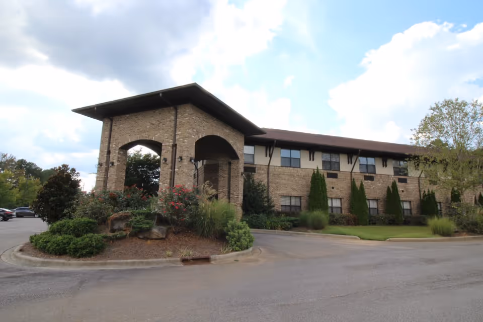 Exterior view of a two-story brick building with a covered entrance supported by large pillars. The building is surrounded by landscaped greenery including bushes, trees, and flowering plants. The sky is partly cloudy.