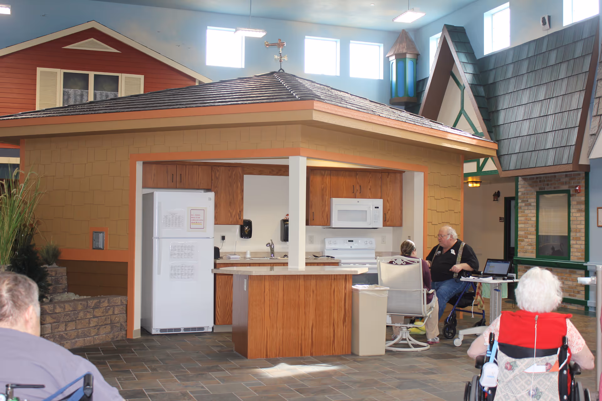 Indoor common area designed to look like a small house with a kitchen setup including a refrigerator, microwave, stove, and wooden cabinets. Several elderly individuals are seated nearby, some in wheelchairs, engaging in conversation. The space has high windows and a bright, welcoming atmosphere with house-like structures in the background.