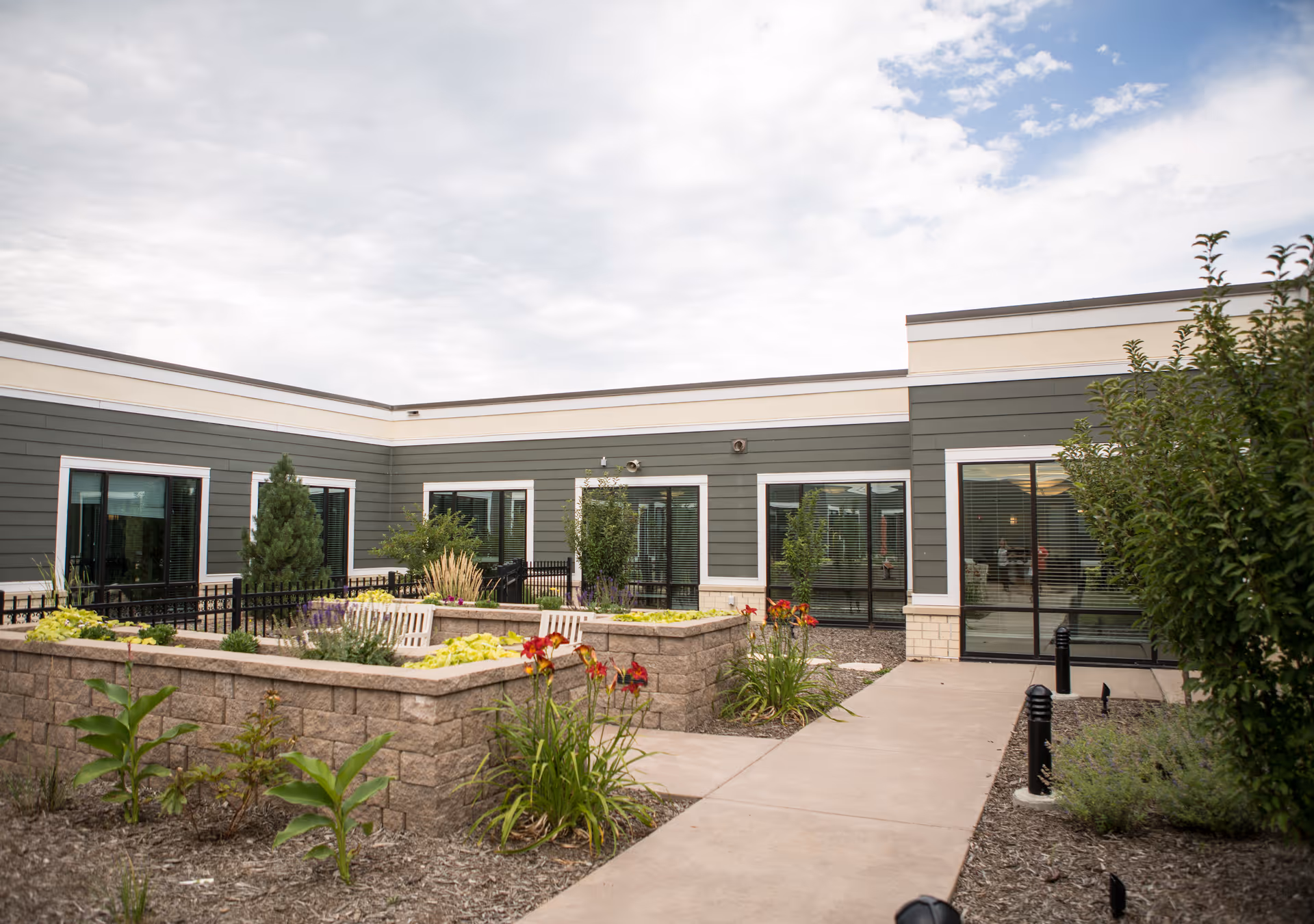 Outdoor courtyard area of a senior living facility with raised garden beds containing various plants and flowers. The courtyard is surrounded by a single-story building with large windows and a paved walkway leading through the space. The sky is partly cloudy.
