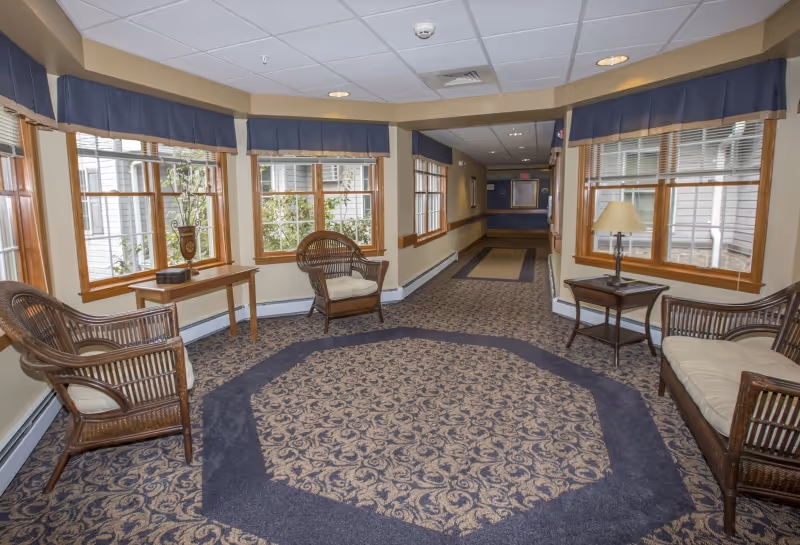 A carpeted hallway with patterned carpet and several wicker chairs with cushions arranged along the walls. There are multiple large windows with blue valances letting in natural light. A small table with a lamp and another table with a decorative vase are also visible. The hallway leads to a corridor with a bulletin board at the end.