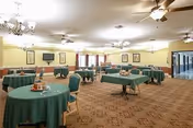 Spacious dining room with round tables covered in green tablecloths, chairs, chandeliers and ceiling fans, and a carpeted floor.