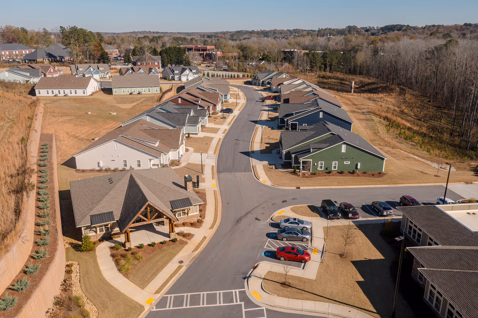 Aerial view of a senior living community with multiple single-story residential buildings arranged along a curved road. There is a parking lot with several cars, landscaped areas, and a wooded area in the background under a clear sky.